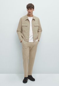 Beige jas met twee voorzakken, gecombineerd met beige broeken en donkere schoenen. De stof heeft een gladde textuur en neutrale tinten.