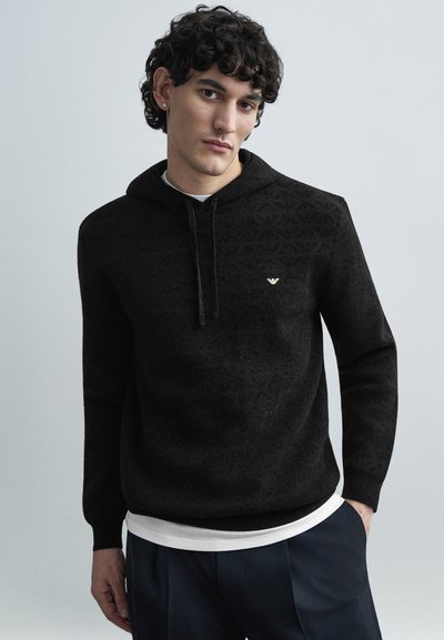 Sweat à capuche noir en tissu texturé, doté d'un cordon de serrage, de poignets côtelés et d'un petit logo doré sur la poitrine. Porté par-dessus une chemise blanche.