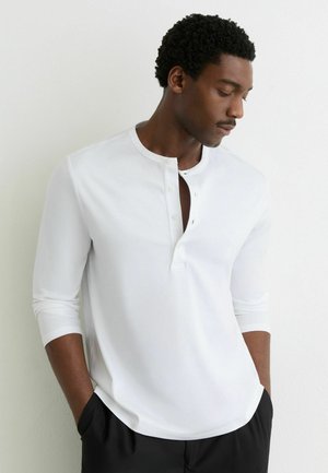 Biela košeľa s dlhým rukávom, okrúhlym výstrihom a štvorradovým gombíkovým zapínaním. Mäkká textúra a uvoľnený strih, v kombinácii s čiernymi nohavicami.