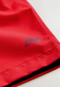 Röda sportshorts av mjukt tyg med en svartaccentuerad fåll och en diskret blå logga på sidan.