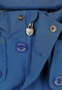 Giacca blu con una texture liscia, dotata di un tirante della zip a forma di orso, bottoni a pressione e un logo ben visibile.
