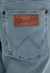 Heledad sinine denimitekk, millel on suur tikitud tagatasku, millel on pruun nahast "Wrangler" silt ja dekoratiivne tikand.