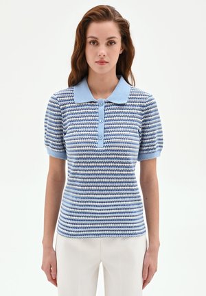 Femme portant un polo en maille à manches courtes avec motif bleu et blanc et boutons, associé à un pantalon blanc, debout devant un fond uni.