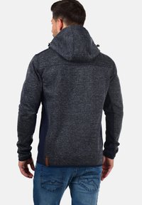 Chaqueta de hombre con capucha en gris oscuro con un acabado de punto texturizado, que presenta paneles laterales en contraste de color marino y un acento de cuero cerca del dobladillo.