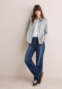 Camicetta a righe verdi e bianche con taschino sul petto, abbinata a pantaloni in denim blu a gamba larga e sneakers nere. Tessuto morbido, design casual.