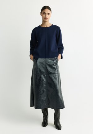Woman standing with hands in pockets wearing navy sweater, black shiny midi skirt, and black knee-high boots against plain white background.