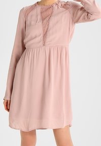 Ljusrosa långärmad klänning med spetsdetaljer vid halsringningen och axlarna, med en lös och flödande silhuett samt texturerade accenter.