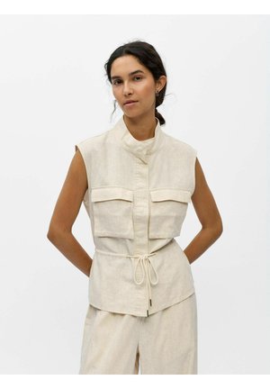 Žena s tmavými vlasmi zviazanými dozadu, oblečená v béžovej ľanovej sleeveless veste s prednými vreckami a zodpovedajúce široké nohavice na bielom pozadí.