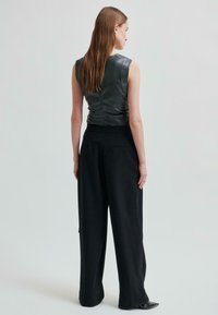 Žena s dlouhými hnědými vlasy, oblečená v bezrukávovém černém koženém topu, širokých černých kalhotách a černých špičatých botách, stojící na jednobarevném pozadí.