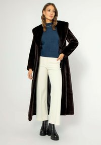 Dlouhý, tmavě hnědý kabát z umělé kožešiny se širokým límcem, spárovaný s modrým svetrem a krémovými kalhotami s širokými nohavicemi. Černé platformové boty dotvářejí celkový vzhled.