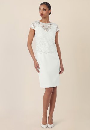 Frau in weißem Spitzen-Oberteil mit kurzen Ärmeln und figurbetontem knielangem Rock, steht mit entspannten Armen da und schaut zur Seite, beige Hintergrund.