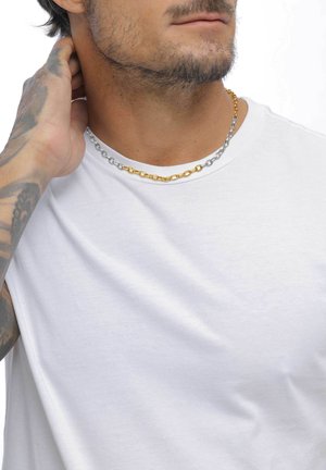 Homme avec une barbe courte portant une chemise blanche et un collier à deux tons, touchant l'arrière de son cou avec une main tatouée.