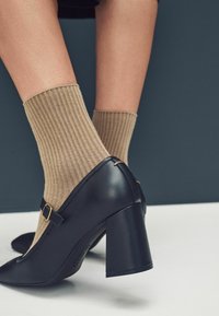 Nohy obuté v béžových žebrovaných ponožkách a černých Mary Jane lodičkách na vysokém podpatku, s kulatou špičkou a blokovým podpatkem, na tmavém a světlém pozadí.