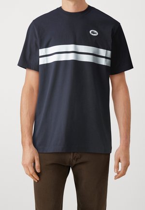 Mann trägt ein schwarzes T-Shirt mit kurzen Ärmeln, zwei horizontalen weißen Streifen und einem kleinen runden Logo auf der Brust.