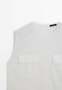 Haut blanc sans manches avec encolure ronde et deux poches avant à rabat avec boutons de chaque côté.