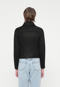 Giacca in suede nera con un design aderente, due tasche laterali e colletto. Indossata sopra jeans chiari a vita alta con una texture sbiadita.