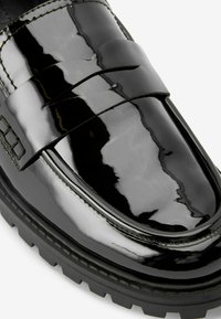 Mocasines de charol negro con un acabado suave y brillante, que presentan una correa horizontal y una suela de goma negra texturizada.