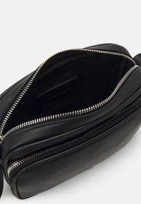 Sort svart skinn crossover-veske med strukturert overflate, utstyrt med en sølvfarget glidelås og et stoffforet indre. Flere rom for organisering.