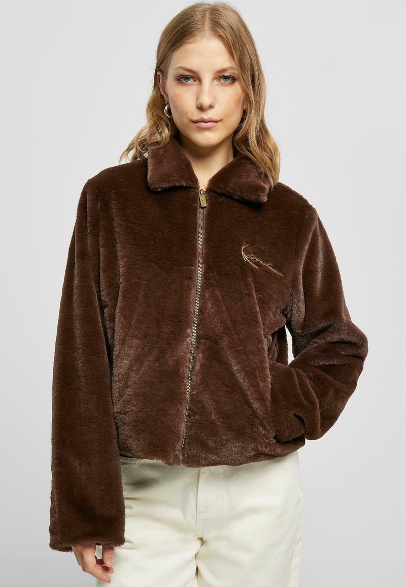 Brun faux fur zip-up jakke med krage, som har lange ermer og et lite brodert design på forsiden. Myk tekstur, kort silhuett.