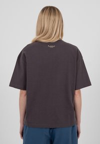 Personne blonde debout, dos à la caméra, portant un T-shirt ample de couleur sombre avec le texte "Interdit" près du col et un pantalon bleu.