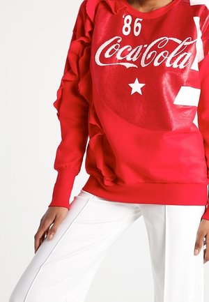 Osoba ubrana w czerwony, długi rękaw z napisem Coca-Cola w białym kolorze i gwiazdą, w połączeniu z białymi spodniami, stojąca na jednolitym tle.