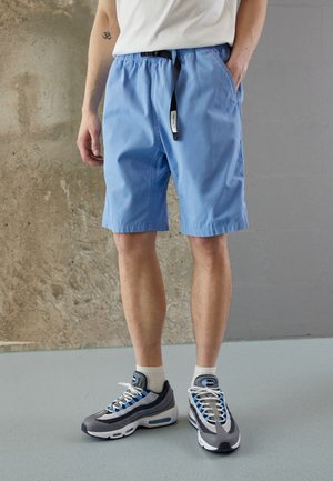 Homme portant un short bleu clair jusqu'aux genoux, des chaussettes blanches et des chaussures de sport grises et bleues, debout contre un mur gris et texturé.