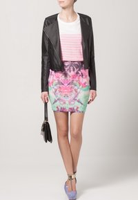 Femme portant une veste en cuir noire, un haut rayé blanc et rose, une jupe florale colorée, tenant un sac à main noir et des escarpins lavande avec des brides dorées autour de la cheville.