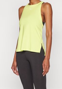Světlé žluté sportovní tílko bez rukávů s kulatým výstřihem, bočními rozparky a logem na hrudi. Doplněno tmavými legínami.
