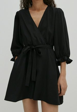 Nainen pukeutuneena mustaan kietaisumekkoon, jossa on pussihihaiset kolmivartiset hihat ja solmittu vyötärövyö, yksinkertaisella taustalla.