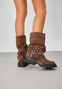 Bottes en daim marron mi-mollet avec des semelles noires épaisses et de multiples œillets argentés, portées sur des jambes nues devant un fond uni.