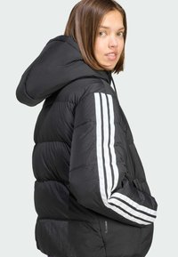 Chaqueta negra acolchada con secciones acolchadas, rayas horizontales blancas en las mangas y capucha; presenta una textura brillante y suave.