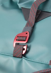 Teal backpack detail featuring a red plastic buckle and gray strap with red accents, smooth texture, and outlined seam detail.