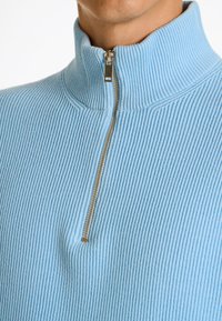 Ljusblå ribbad halvdragkedja med hög krage och metallisk dragkedje-detalj. Mjuk textur med vertikala ränder.