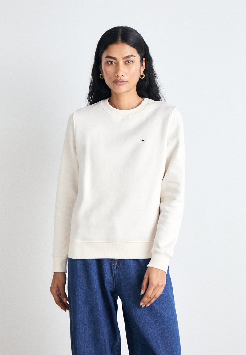 Sweatshirt crème avec un col rond et des poignets côtelés. Présente un petit logo sur la poitrine. Associé à un jean bleu ample.