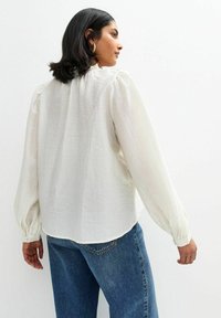 Witte textuurblouse met pofmouwen, ronde halslijn en gathered details, gecombineerd met blauwe denim jeans.