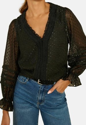 Vrouw draagt een zwarte doorschijnende blouse met polka dots, met kanten rand en pofmouwen, gecombineerd met een blauwe spijkerbroek en een zwarte riem.