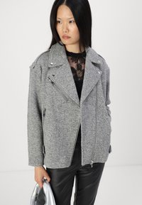 Veste de moto en mélange de laine texturée gris, avec fermeture éclair à l'avant, épaulettes et poches latérales, assortie à un haut en dentelle noire transparente et à un pantalon en cuir.