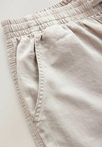 Pantalon en tissu léger beige avec taille élastique et poche latérale, montrant des coutures détaillées et la texture.