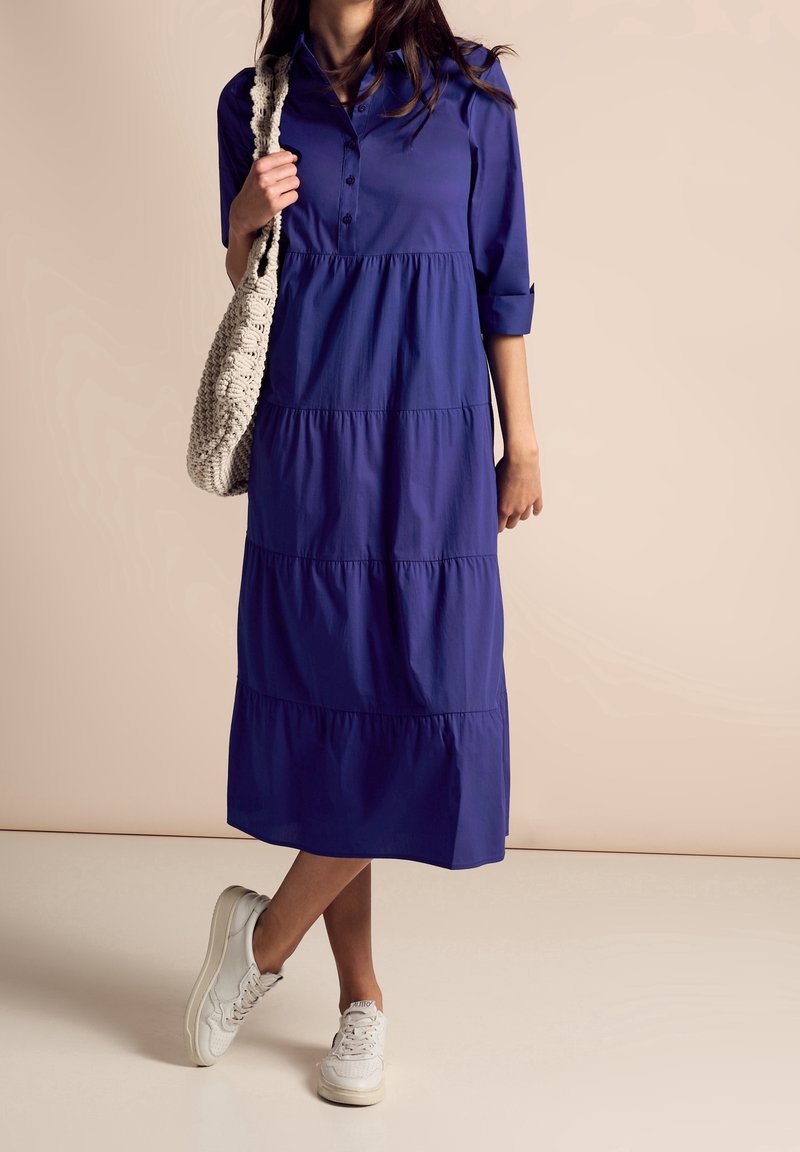 Femme portant une robe midi bleue à volants, des baskets blanches et tenant un sac à épaule beige en tricot.