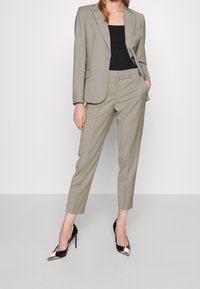 Kvinna i en ljusgrå skräddarsydd kostym med korta byxor, svart topp och svarta spetsiga högklackade skor med silverdetaljer.