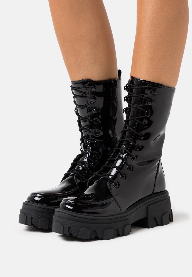 Bottes de combat à lacets en cuir verni noir brillant avec semelles à crampons épaisses portées sur des jambes nues sur un fond blanc.