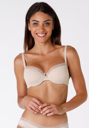 Brasiletta imbottita beige con bordi scallop e spalline regolabili. Caratterizzata da una finitura liscia e chiusura frontale. Il modello assume una posa rilassata.
