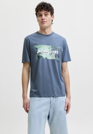 Jeune homme aux cheveux courts et bouclés porte un T-shirt bleu à motif tropical et un jean bleu clair, debout devant un fond blanc uni.