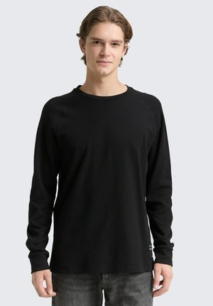 Giovane uomo con capelli castano chiaro che indossa una maglietta nera a maniche lunghe con scollo tondo e jeans grigi, in piedi davanti a uno sfondo chiaro e semplice.