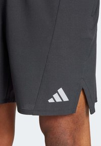 Svarta atletiska shorts med texturerad tyg, sidofickor och en vit Adidas-logotyp på nederkanten.