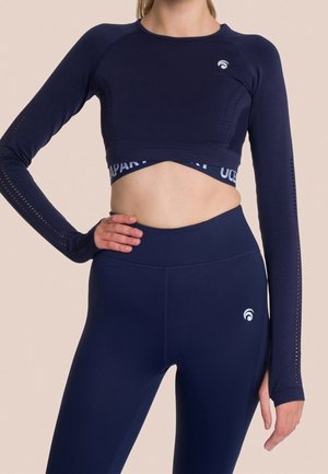 Kvinde iført navy langærmet crop top og højtaljede leggings med subtile logodetaljer på beige baggrund.
