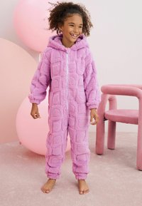 Combinaison violette duveteuse avec une capuche, présentant un motif carré texturé, des poignets élastiques et une fermeture éclair à l'avant. Tissu doux et pelucheux visible.