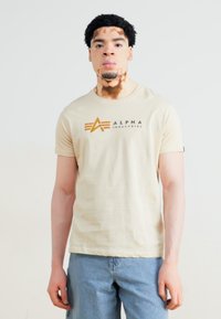 Bežīgs kokvilnas T-krekls ar apaļu kakla izgriezumu un īsām piedurknēm; rotāts ar tumši oranžu "ALPHA INDUSTRIES" logotipu ar grafiskām detaļām.