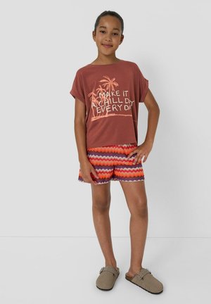 Dziewczyna stojąca z ręką na biodrze, ubrana w rdzawe t-shirt z nadrukiem palm i tekstem, krótkie spodenki w zygzakowaty wzór oraz beżowe buty wsuwane.