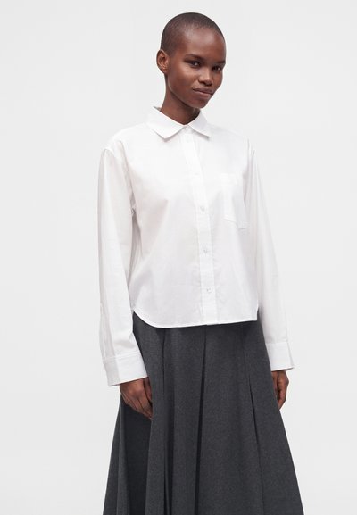 Femme aux cheveux courts portant une chemise blanche boutonnée à manches longues et une jupe plissée gris foncé sur un fond uni.